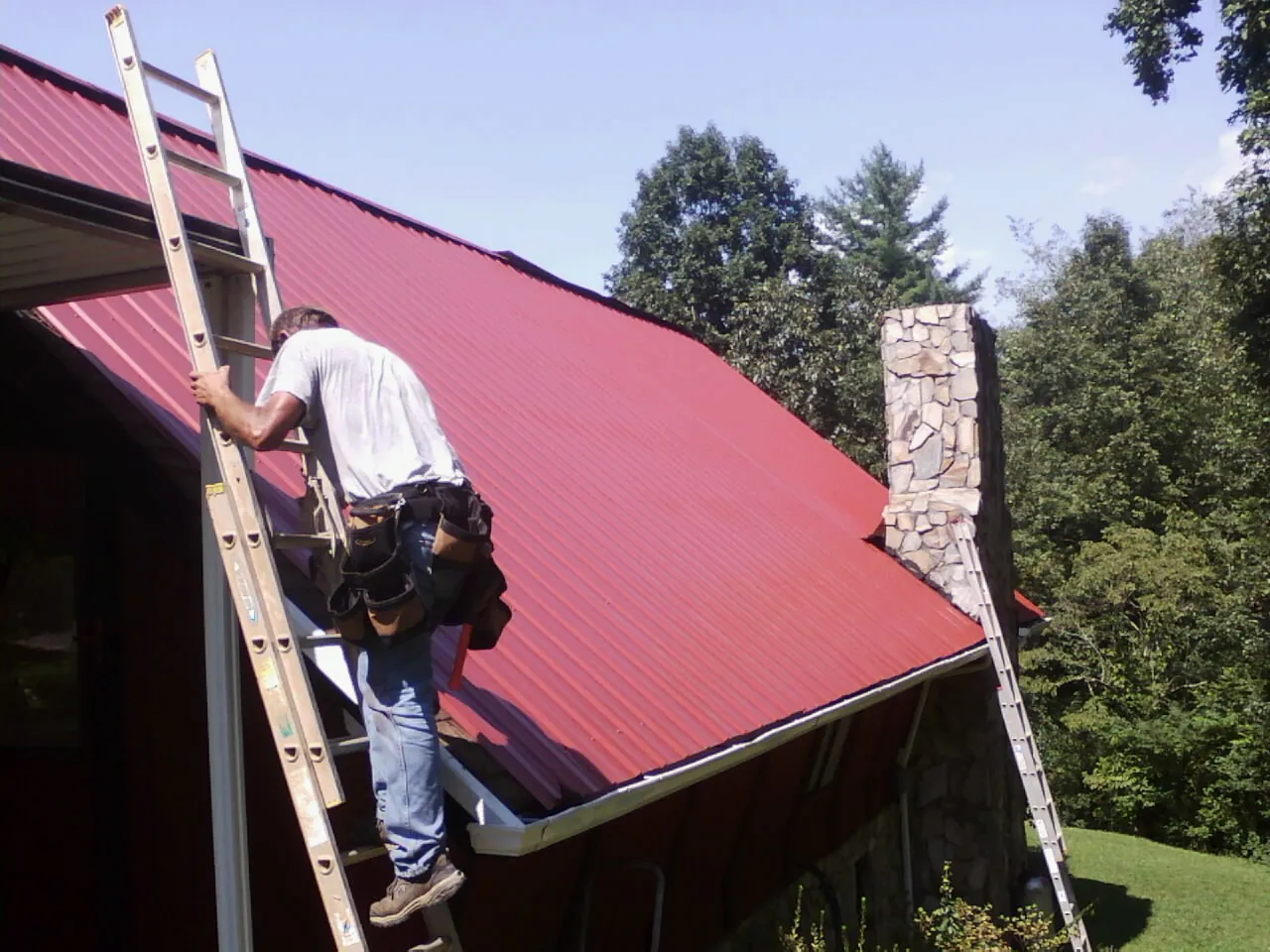 Roofer on a ladder beside a red metal roof and stone chimney — Soffit & Fascia Repair by Roofing Brothers in Estell Manor
